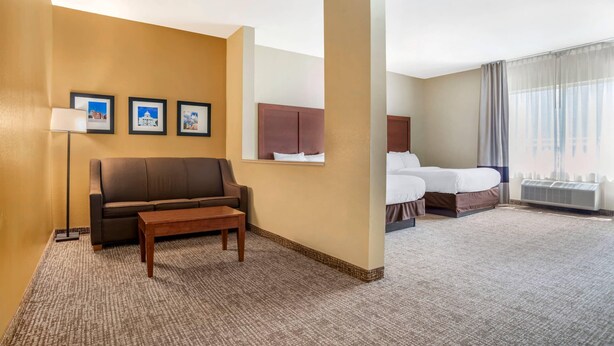 Quarto de hotel com um sofá marrom, uma mesa pequena, uma cama com edredom branco e uma grande janela com cortinas.
