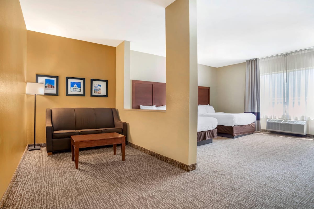 Una habitación de hotel con un sofá marrón, una mesita, una cama con edredón blanco y un ventanal con cortinas.