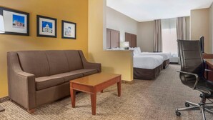 Quarto de hotel com um sofá marrom, uma mesa de centro de madeira, duas camas de solteiro, uma escrivaninha com cadeira e quadros nas paredes.