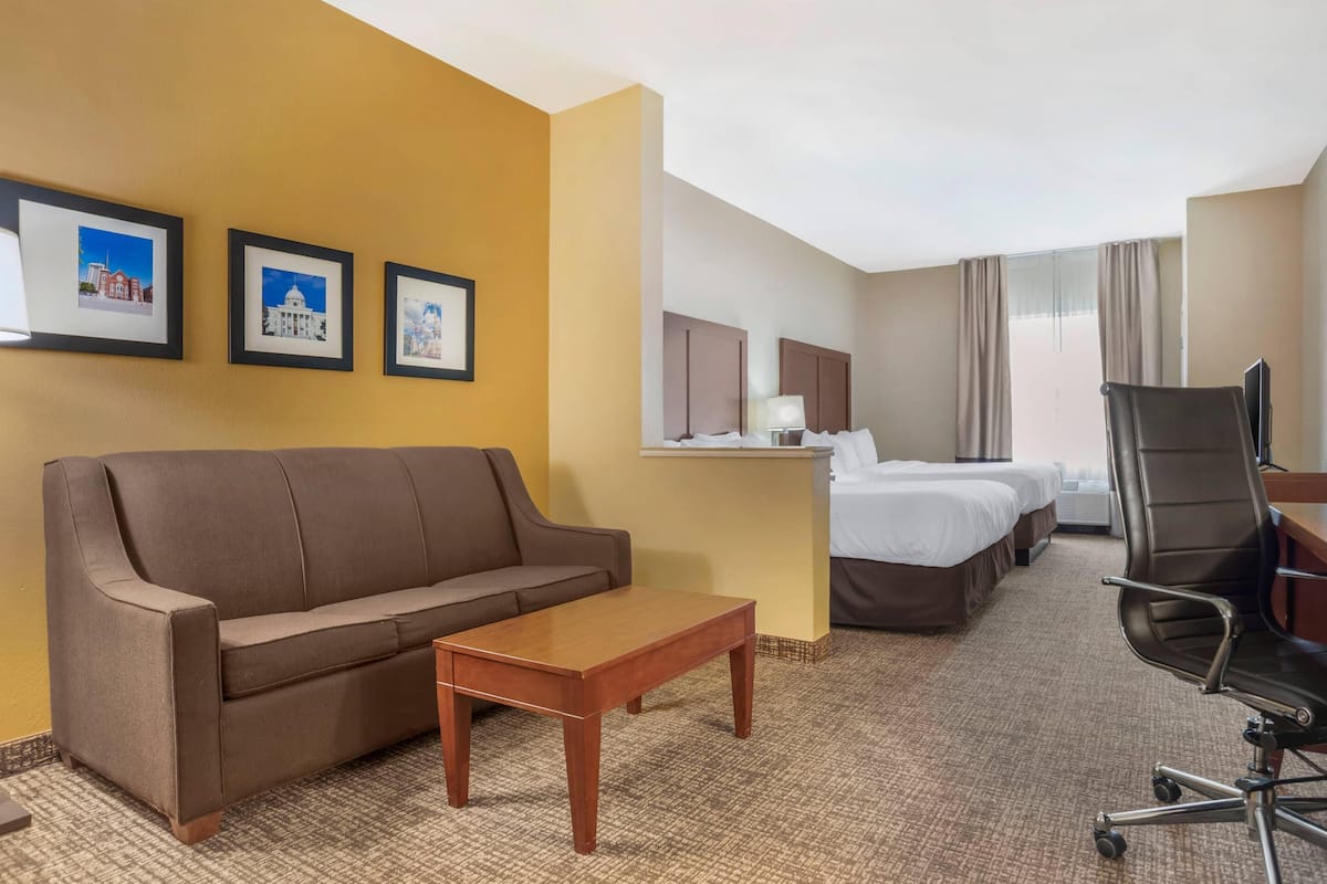 Una habitación de hotel con un sofá marrón, una mesa de centro de madera, dos camas individuales, un escritorio con una silla y cuadros en la pared.