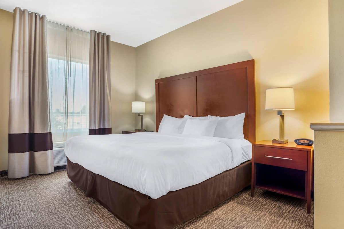 Habitación de hotel con una cama grande, cabecera de madera, mesita de noche con lámpara y vista a la ciudad por la ventana.