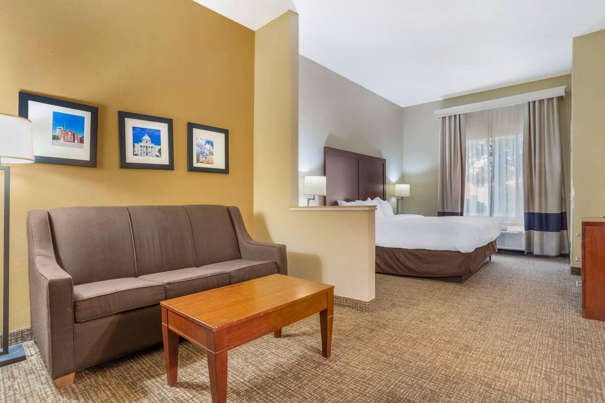 Habitación de hotel con un sofá marrón, una mesa de madera, una cama con colcha blanca, un escritorio con computadora y cuadros en la pared.