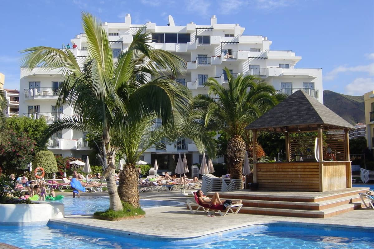 A pool area with lounge chairs, palm trees, and a building with balconies in the background.