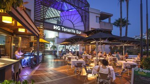 An outdoor dining area with tables and chairs, a piano, and a sign for "HG Tenerife Sur". - Apartamentos HG Tenerife Sur (Arona)