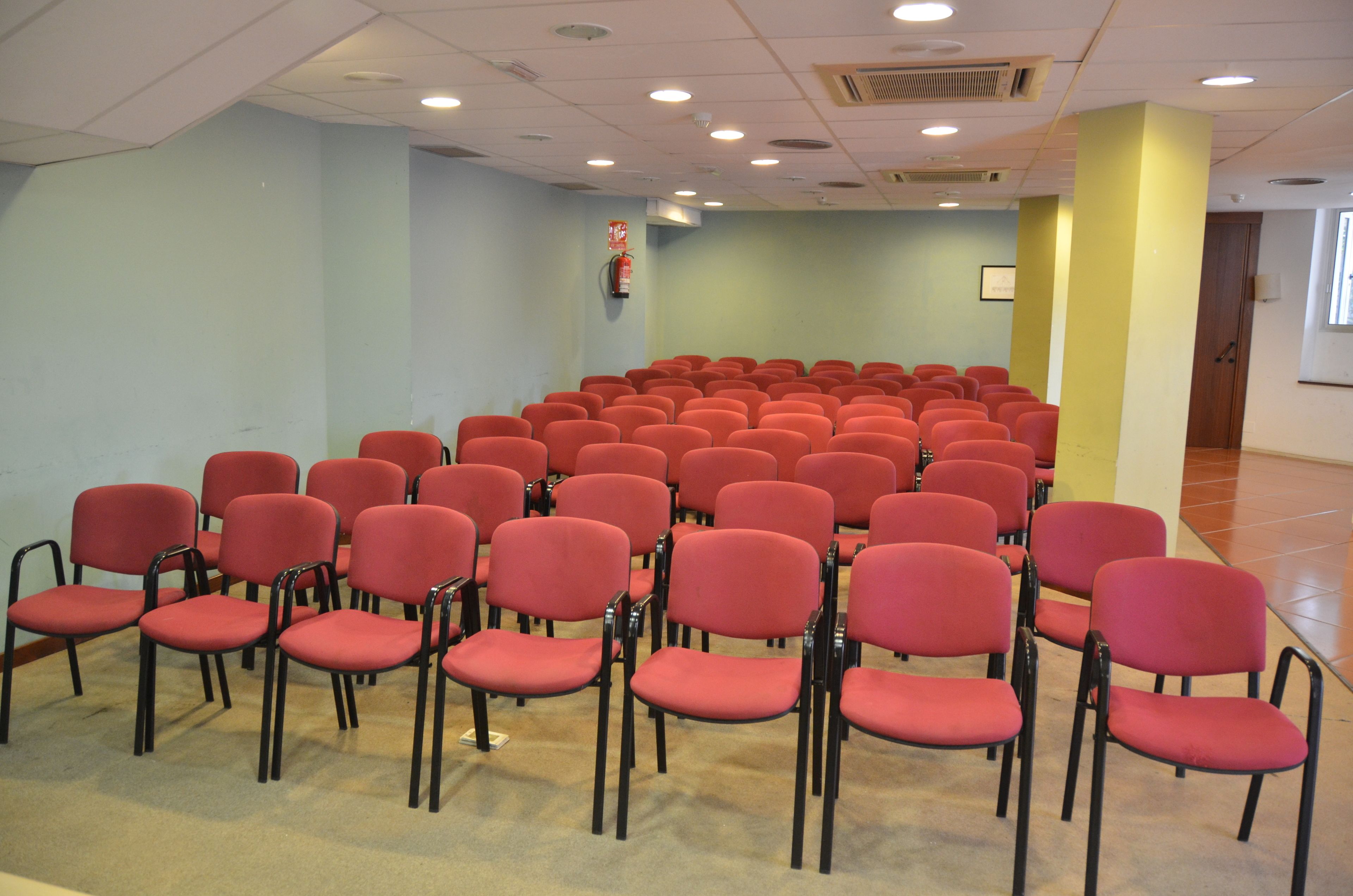 Une salle de conférence avec des rangées de chaises rouges et un extincteur accroché au mur.