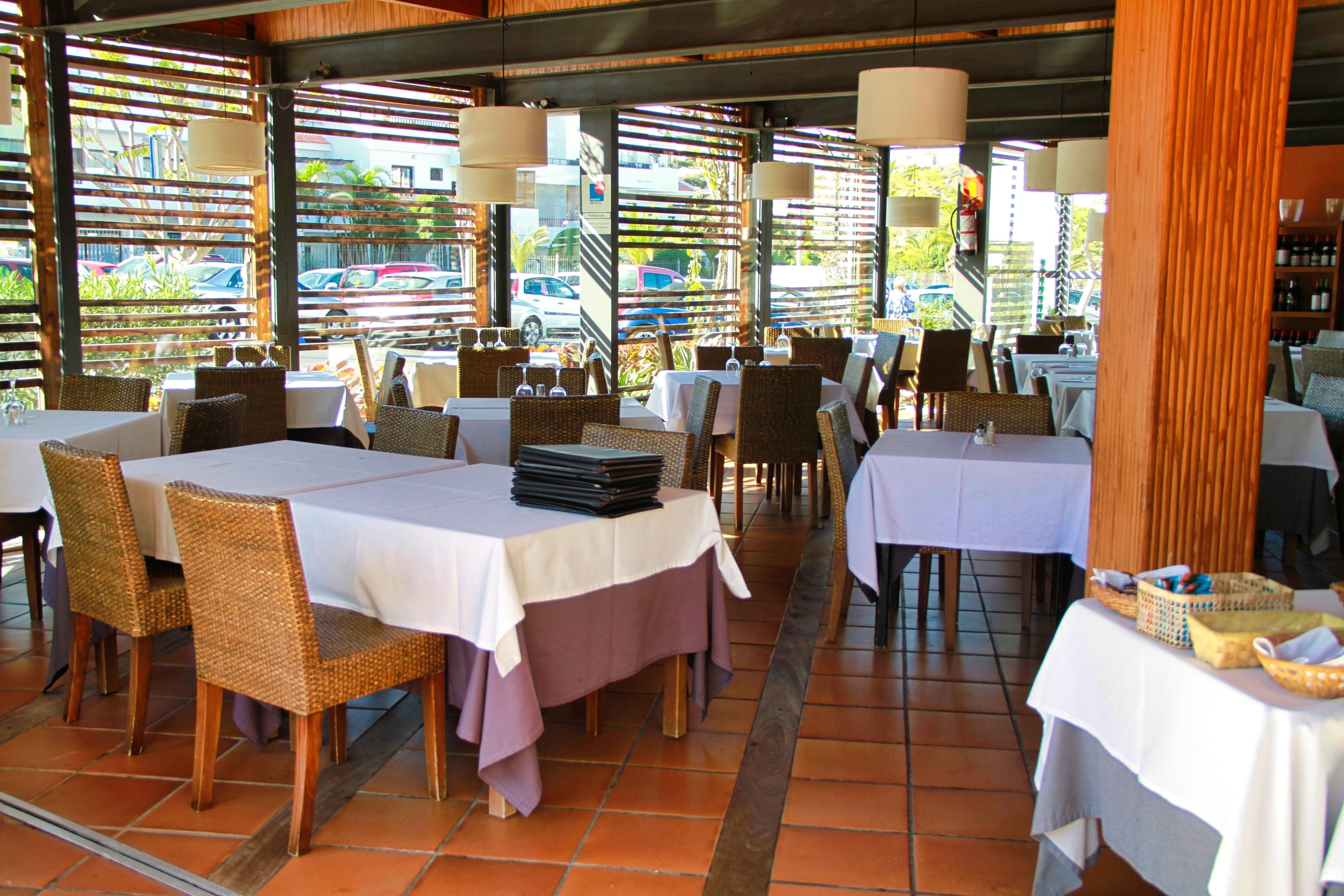 An outdoor restaurant with tables and wicker chairs, covered by a roof with wooden slats and hanging lamps.