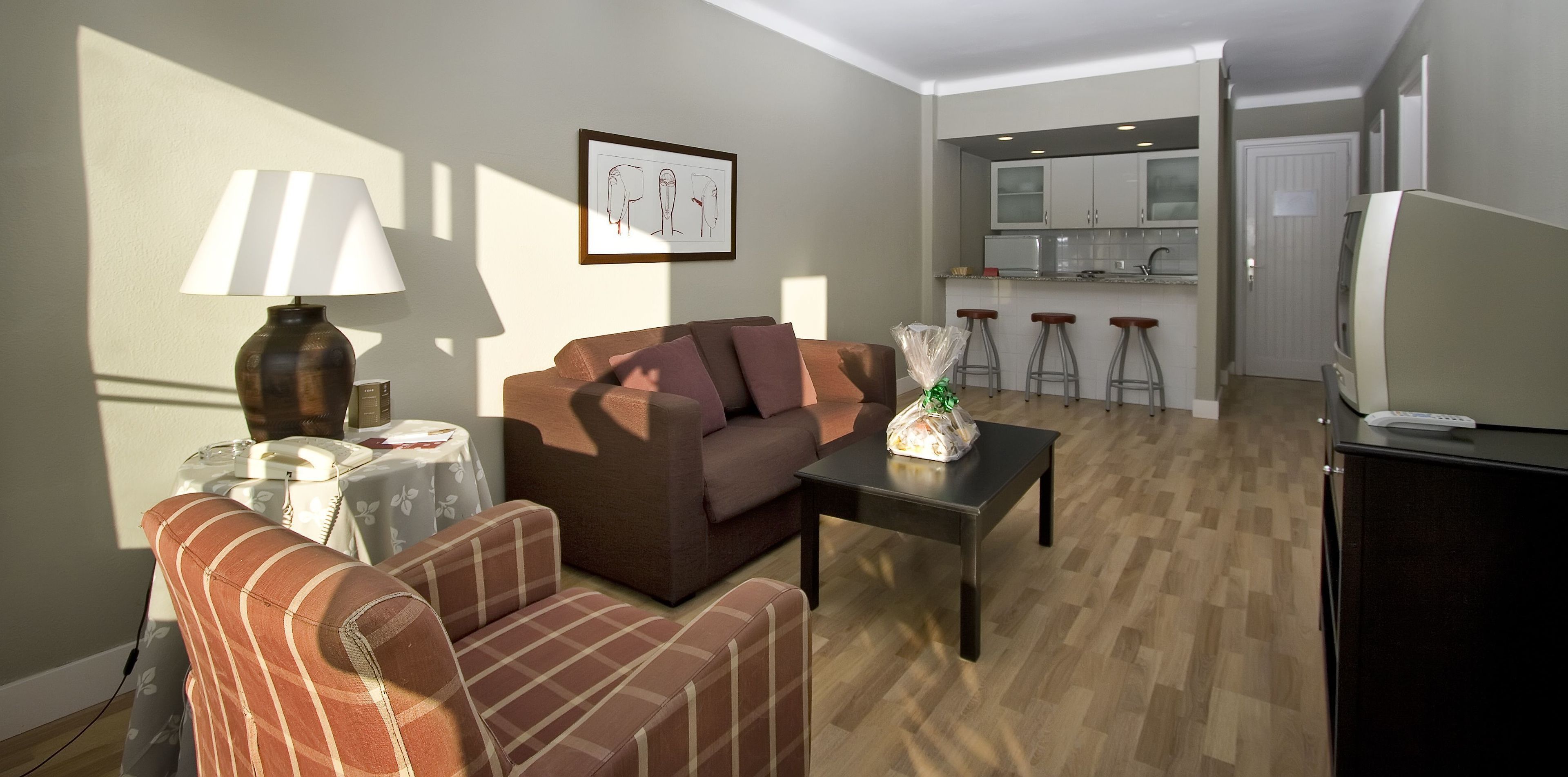 A living room with a sofa, a coffee table, and a lamp. There is a kitchen in the background with a microwave and a refrigerator.