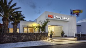 A modern hotel entrance with a stone wall, palm trees, and a sign reading "HG Lomo Blanco". - Apartamentos HG Lomo Blanco (Tías)