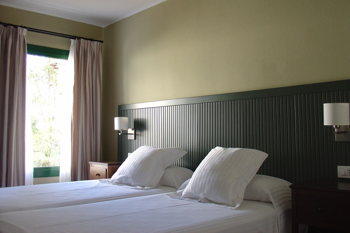 A hotel room with two beds, a nightstand, a lamp, and a window with curtains.