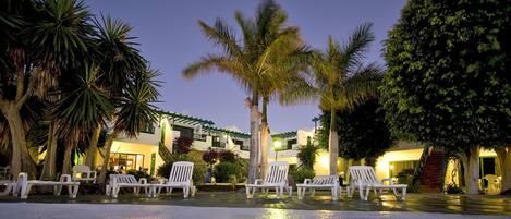 Un resort con sillas de descanso blancas dispuestas en filas, palmeras y un edificio con múltiples balcones y ventanas.