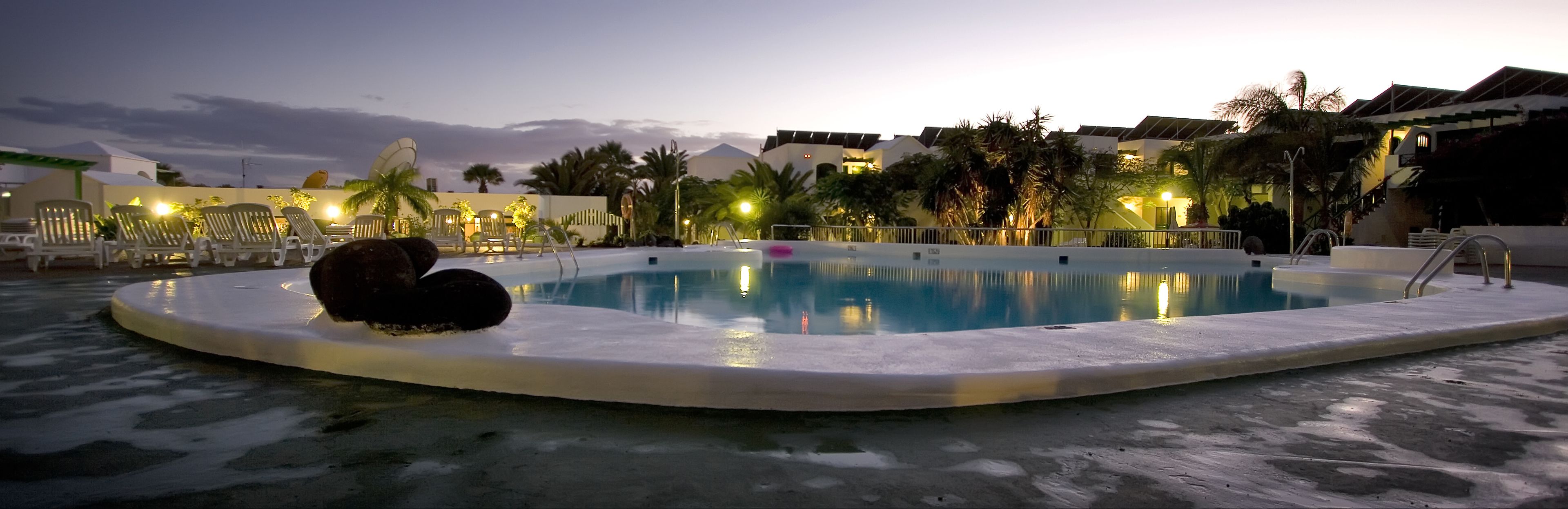 A swimming pool with lounge chairs and palm trees, surrounded by buildings with lights on.