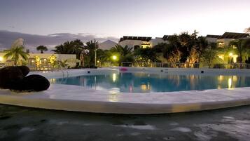 Una piscina con sillones de descanso y palmeras, rodeada de edificios con luces encendidas.