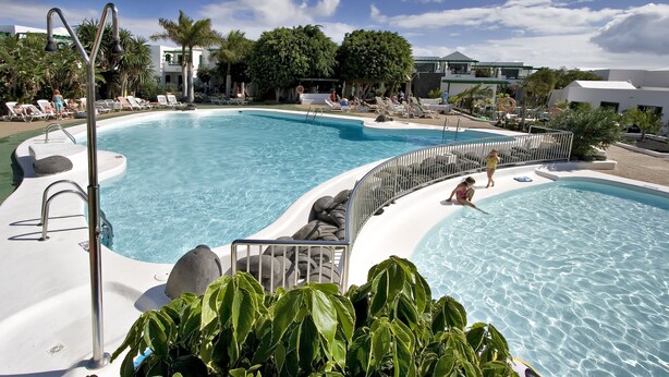 A pool area with multiple sections, including a main pool and a smaller kiddie pool. There are lounge chairs and umbrellas around the pool. - Apartamentos HG Lomo Blanco (Tías)