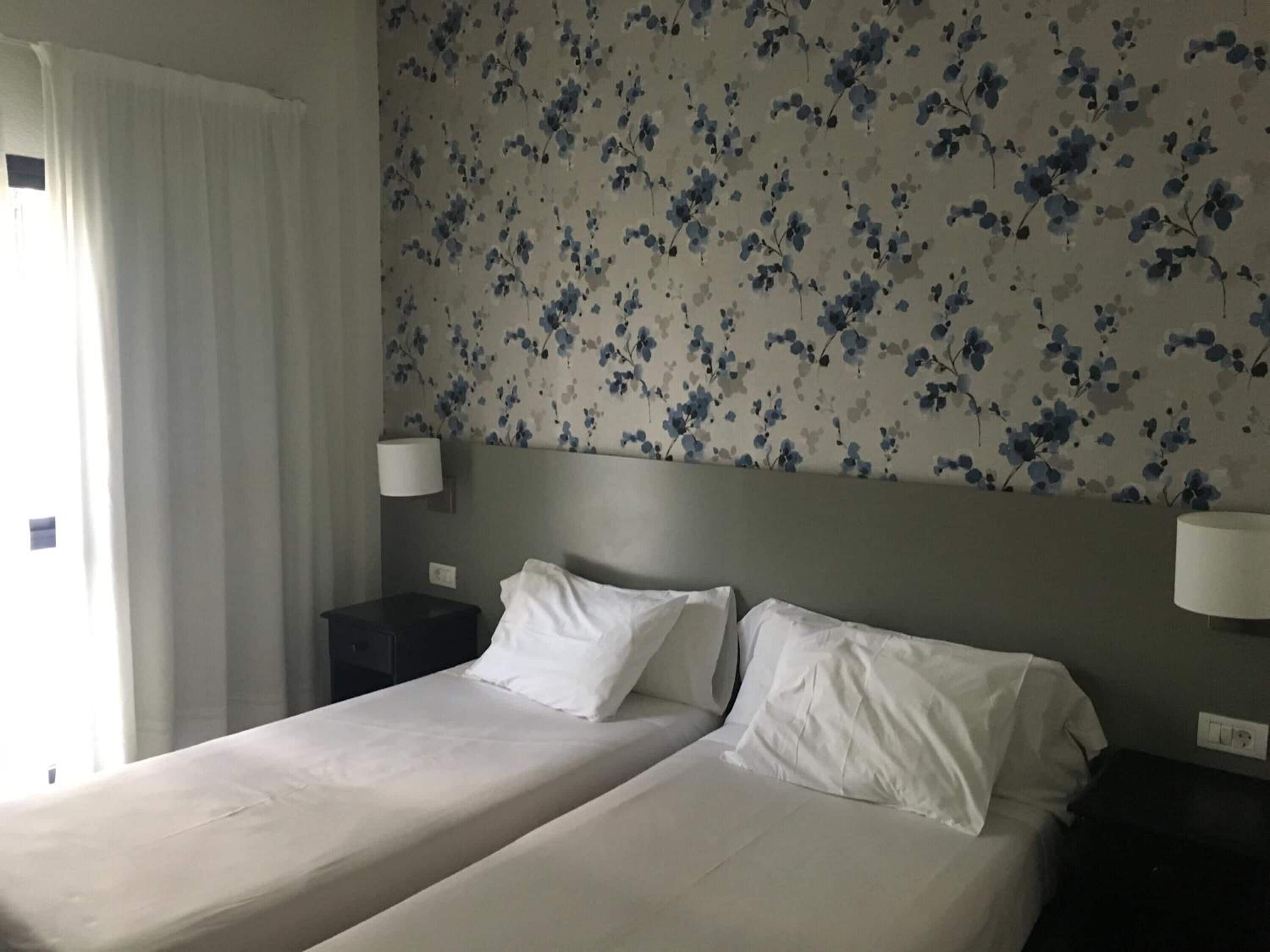 A hotel room with two beds, a floral wallpaper, and two bedside lamps.
