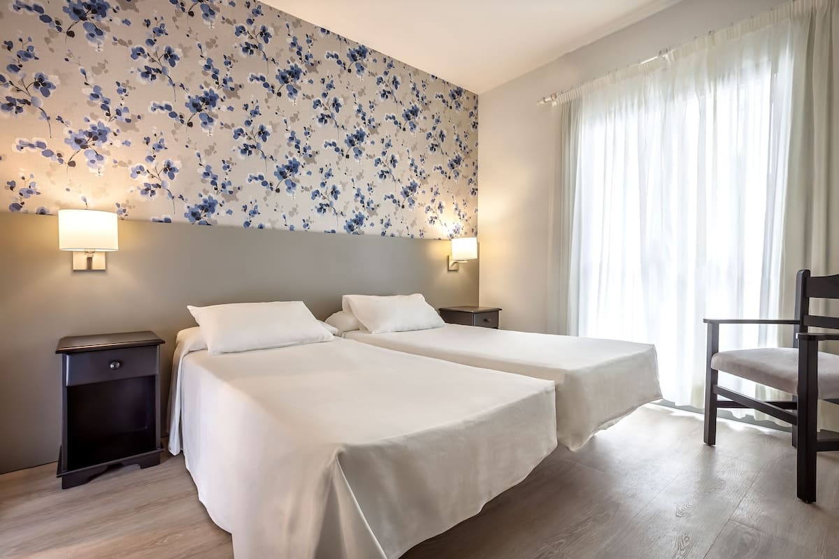 A hotel room with two beds, a nightstand, a chair, and a floral wallpaper.