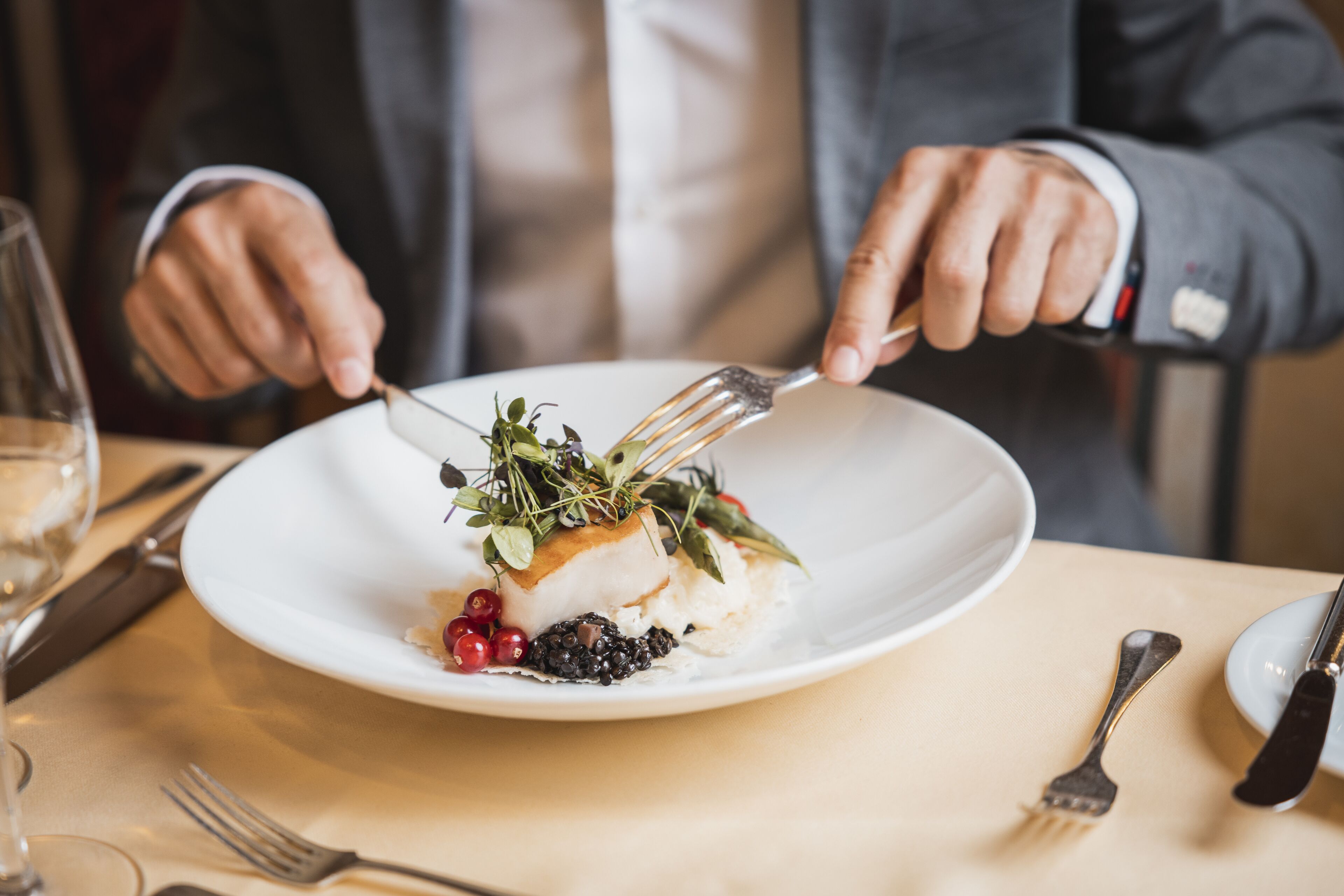 Una persona trajeada cenando en una mesa, con un plato blanco de comida, un tenedor y un cuchillo.