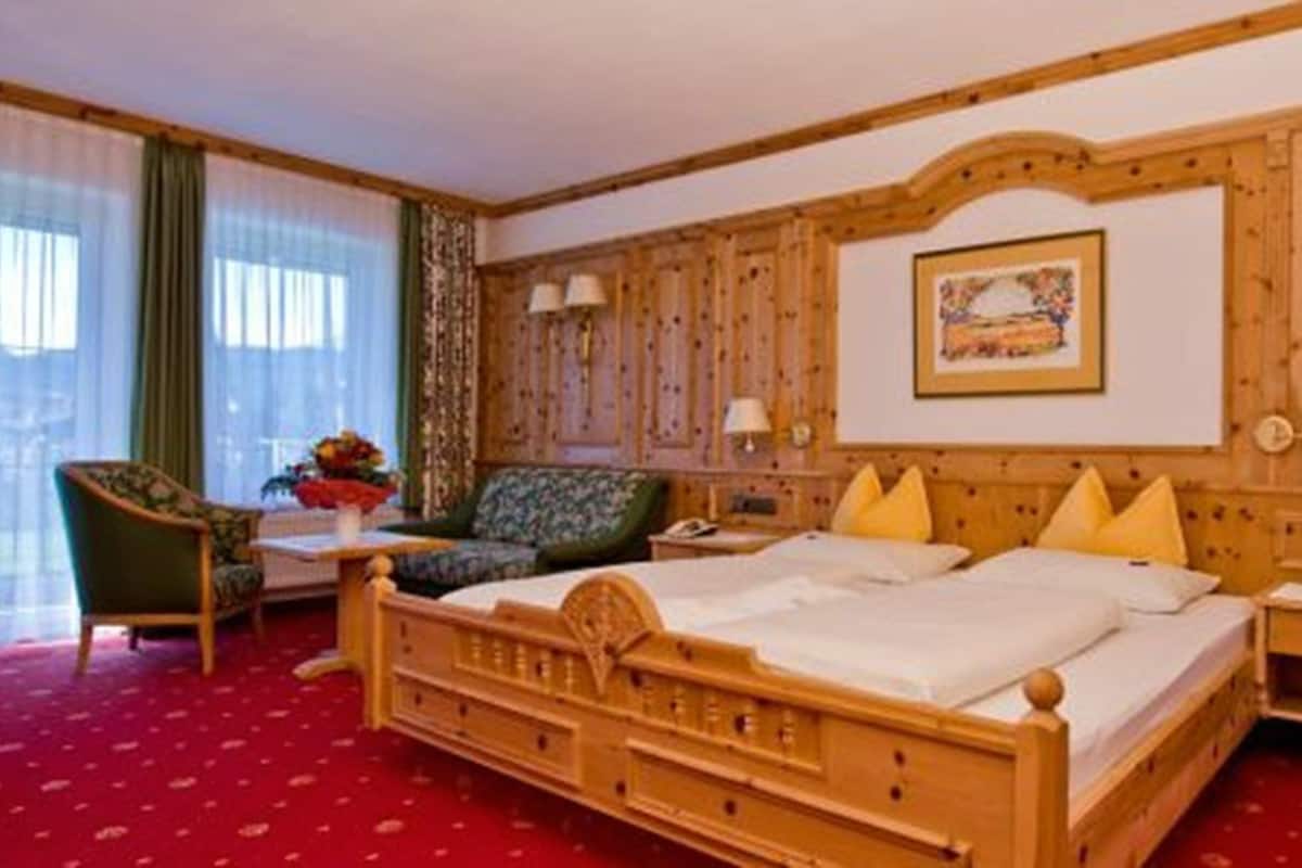 A hotel room with a wooden bed, a green sofa, a small table with a vase, and a window with sheer curtains.