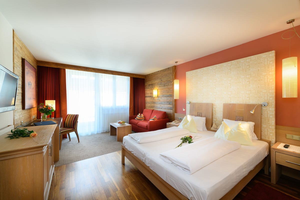 A hotel room with a large bed, a desk with a television, a chair, a small table, and a wall-mounted lamp.