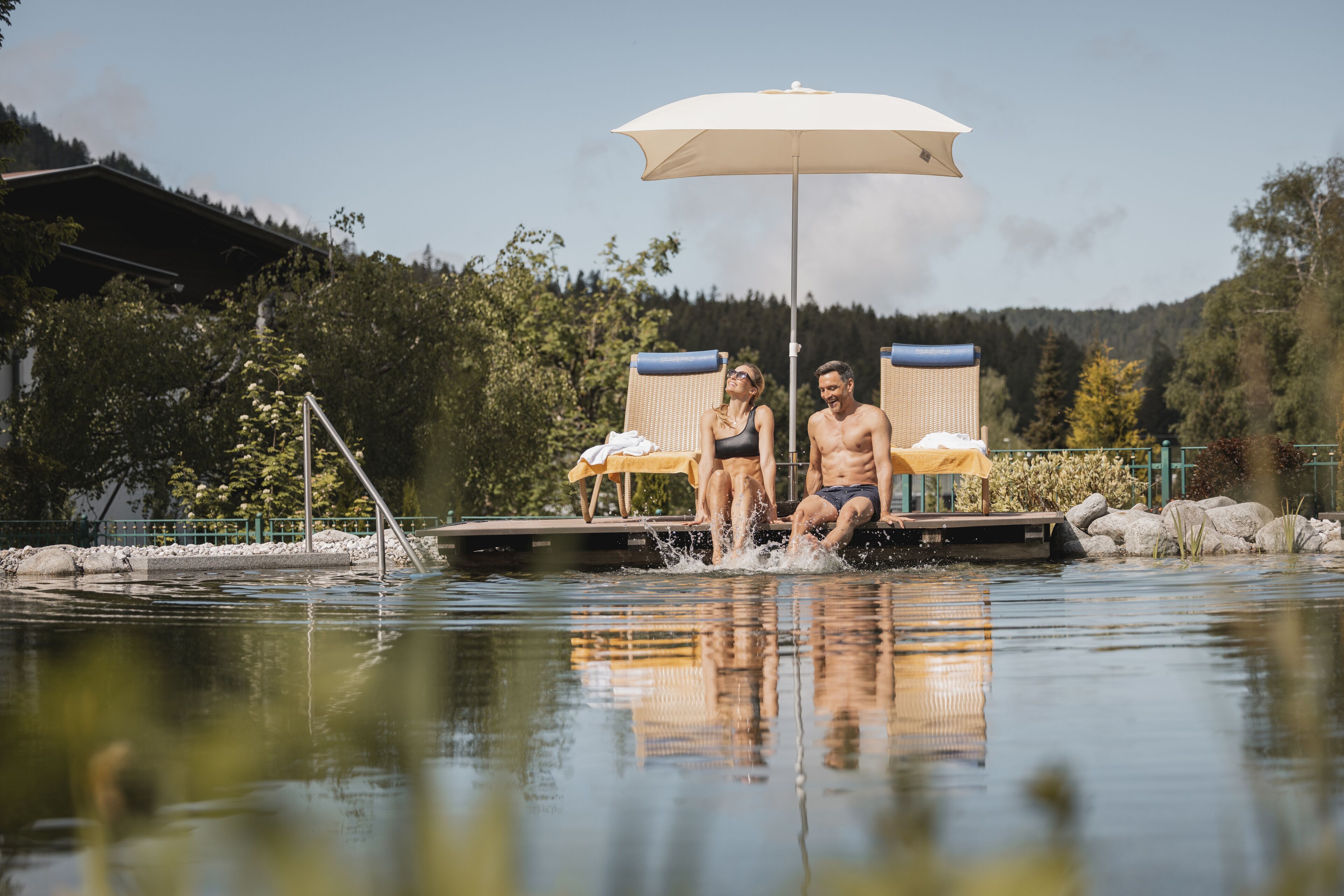 Ein Paar sitzt an einer Anlegestelle an einem stillen See, ein Sonnenschirm spendet Schatten.