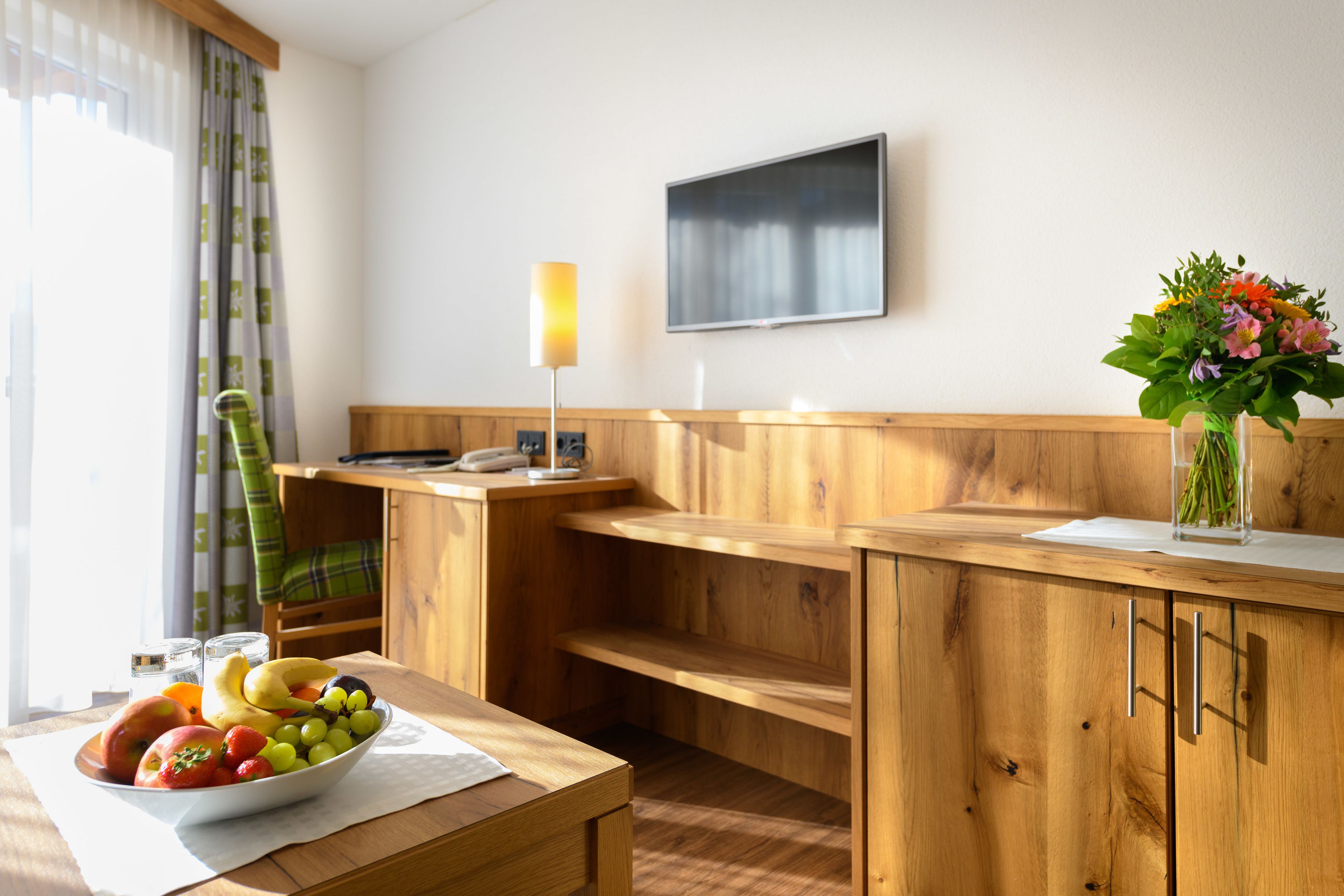 Una habitación de madera con una mesa de comedor, un frutero, un jarrón con flores, un televisor de pantalla plana y una ventana con cortinas.