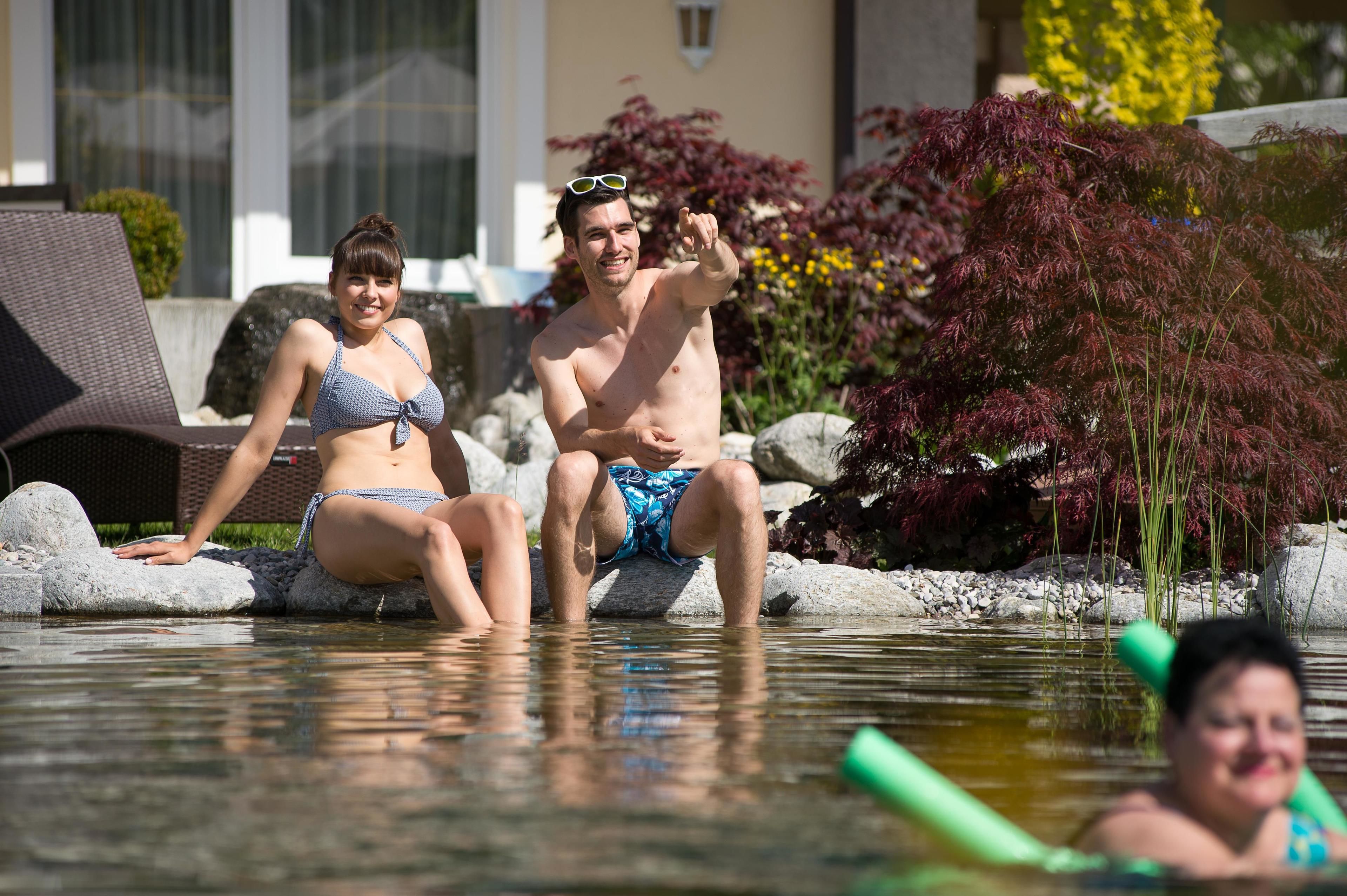 Two people sitting on rocks in a pool area with a building and plants in the background.