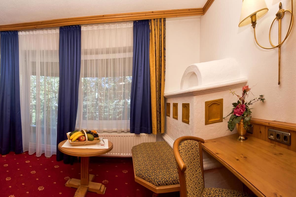 A hotel room with a wooden desk, a television, a small round table with fruit, a leopard print chair, a radiator, and a window with blue curtains.