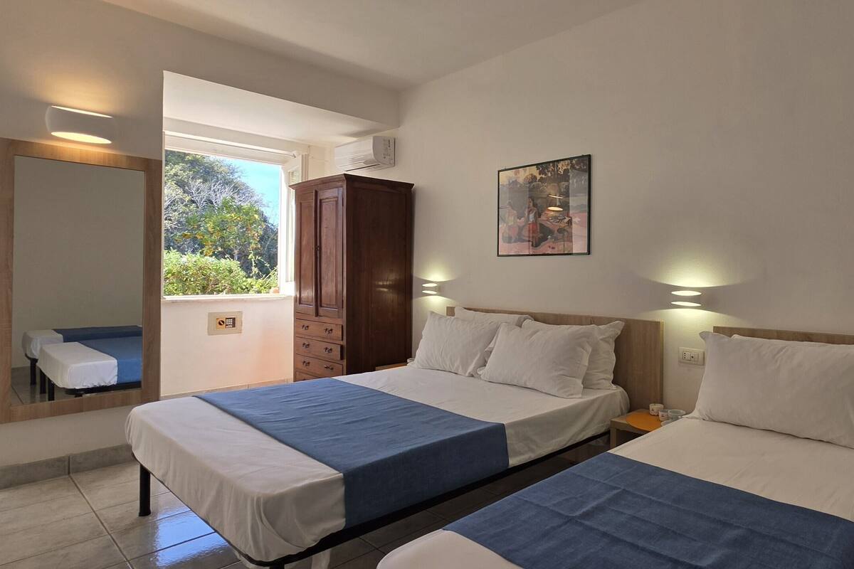 Hotelkamer met twee bedden, een klein tafeltje, een stoel, een schilderij aan de muur en een raam met uitzicht op groen.