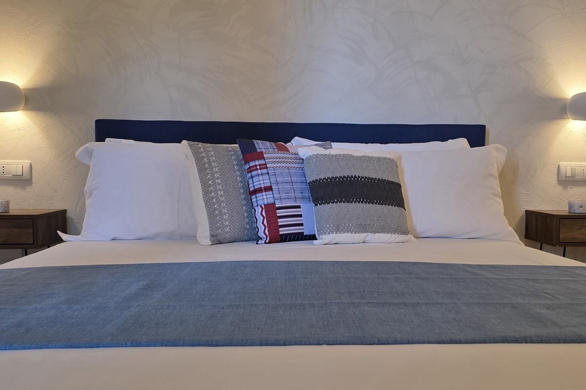 Een opgemaakt bed met een blauwe deken, meerdere kussens en twee nachtkastjes met lampen.