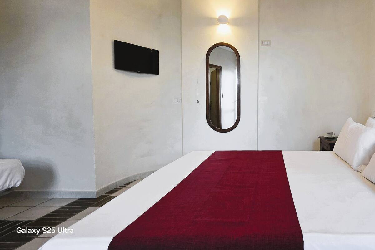 Een net opgemaakt bed met witte lakens en een rode deken, een spiegel aan de muur en een klein nachtkastje met een kopje.