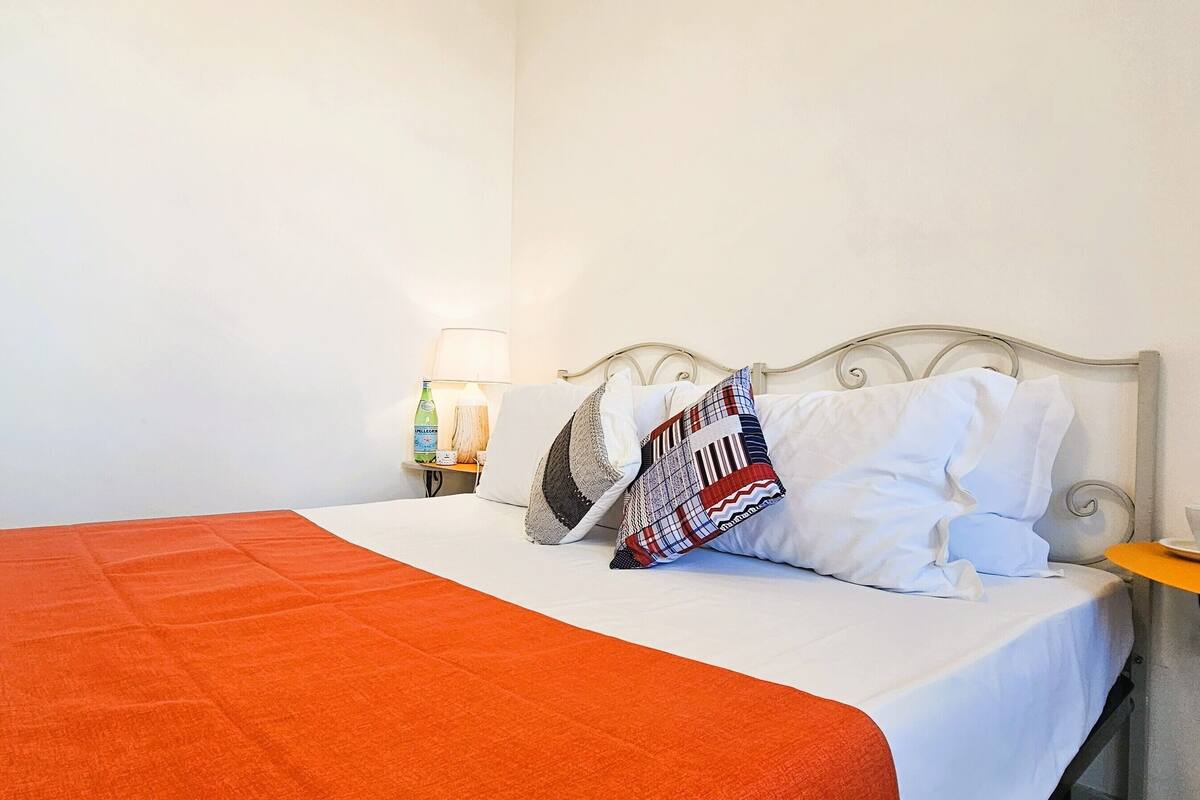 Een net opgemaakt bed met een oranje deken, een gestreept kussen en een wit kussen. Er staat een klein tafeltje met een kop en schotel, en een lamp aan de muur.