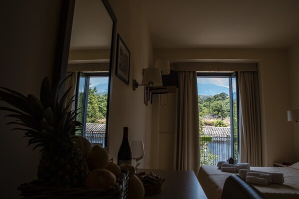 A hotel room with a bed, a dining table with a pineapple and fruit bowl, a wine bottle, and a view of a mountainous landscape through large windows. - Hotel Milomax (Milo)