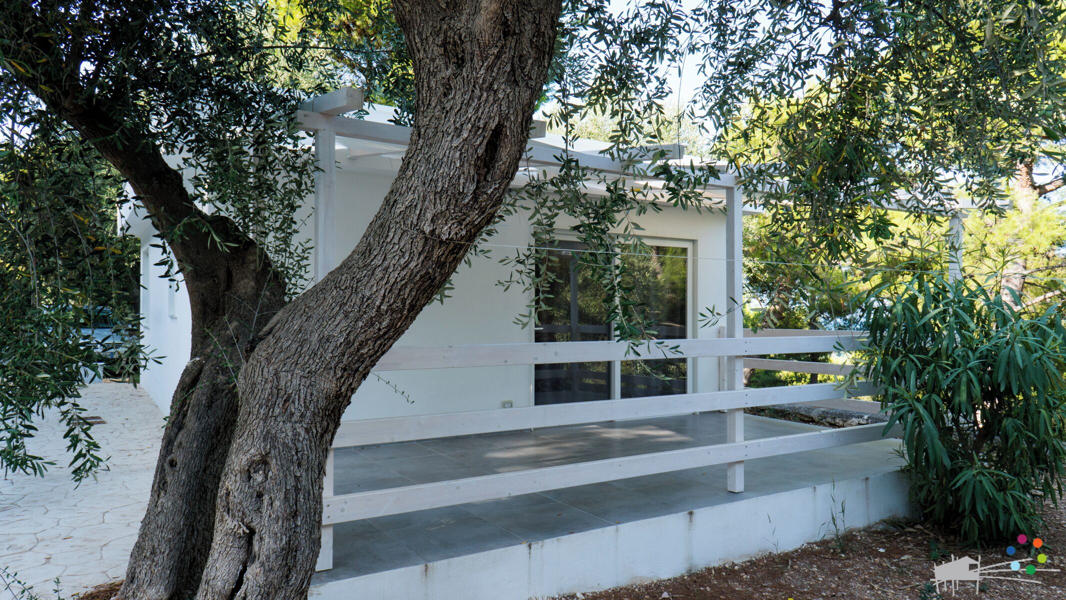 A white building with a wooden deck and a large tree in front.