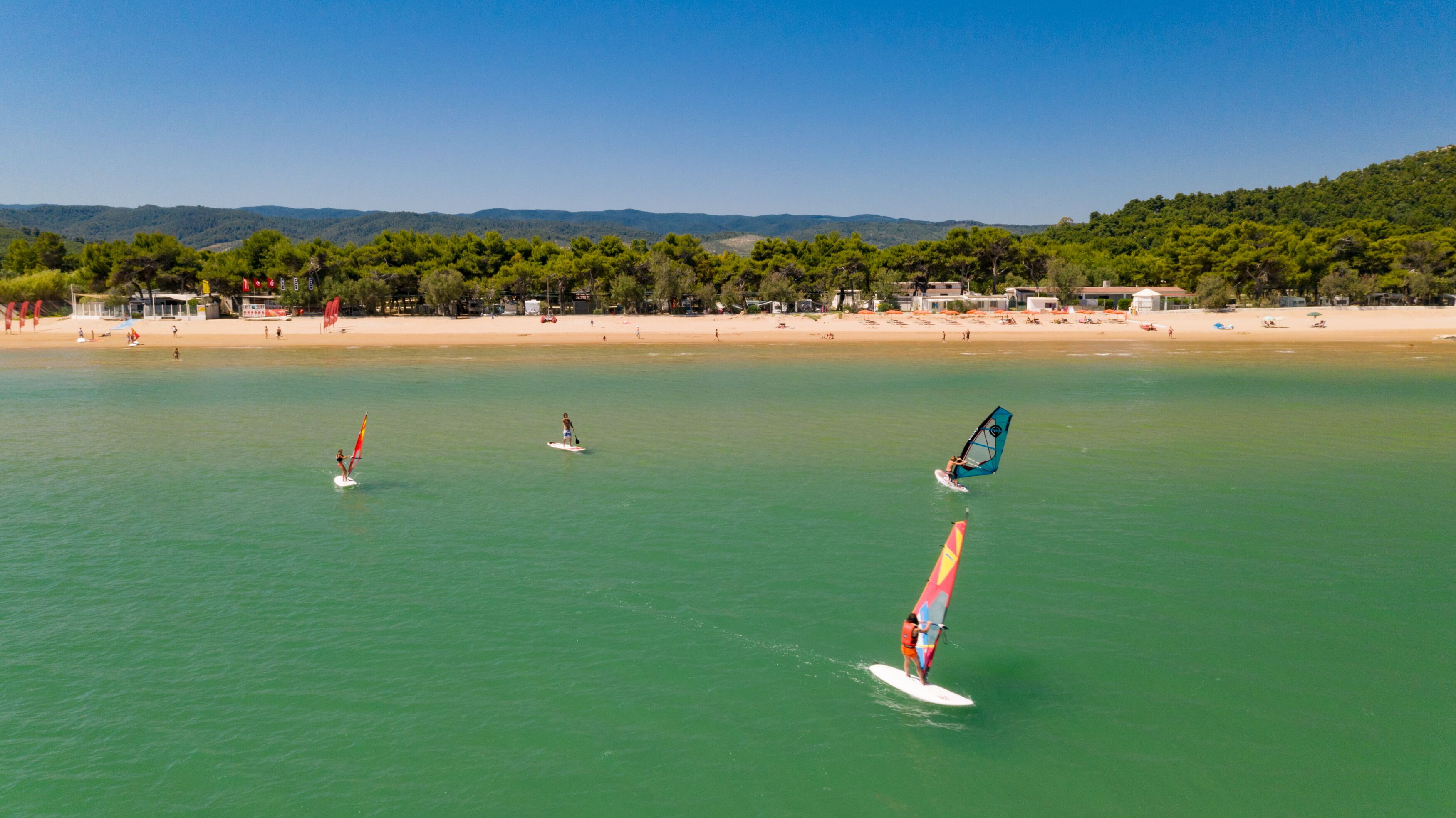Une plage où l’on pratique la planche à voile, avec un ciel dégagé et une colline boisée en arrière-plan.