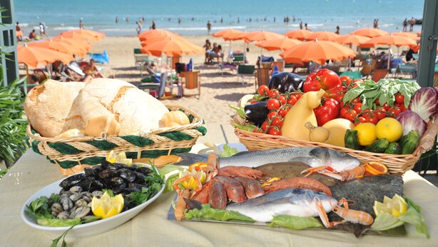 A beachside table with seafood and bread baskets, beach umbrellas, and a view of the ocean.