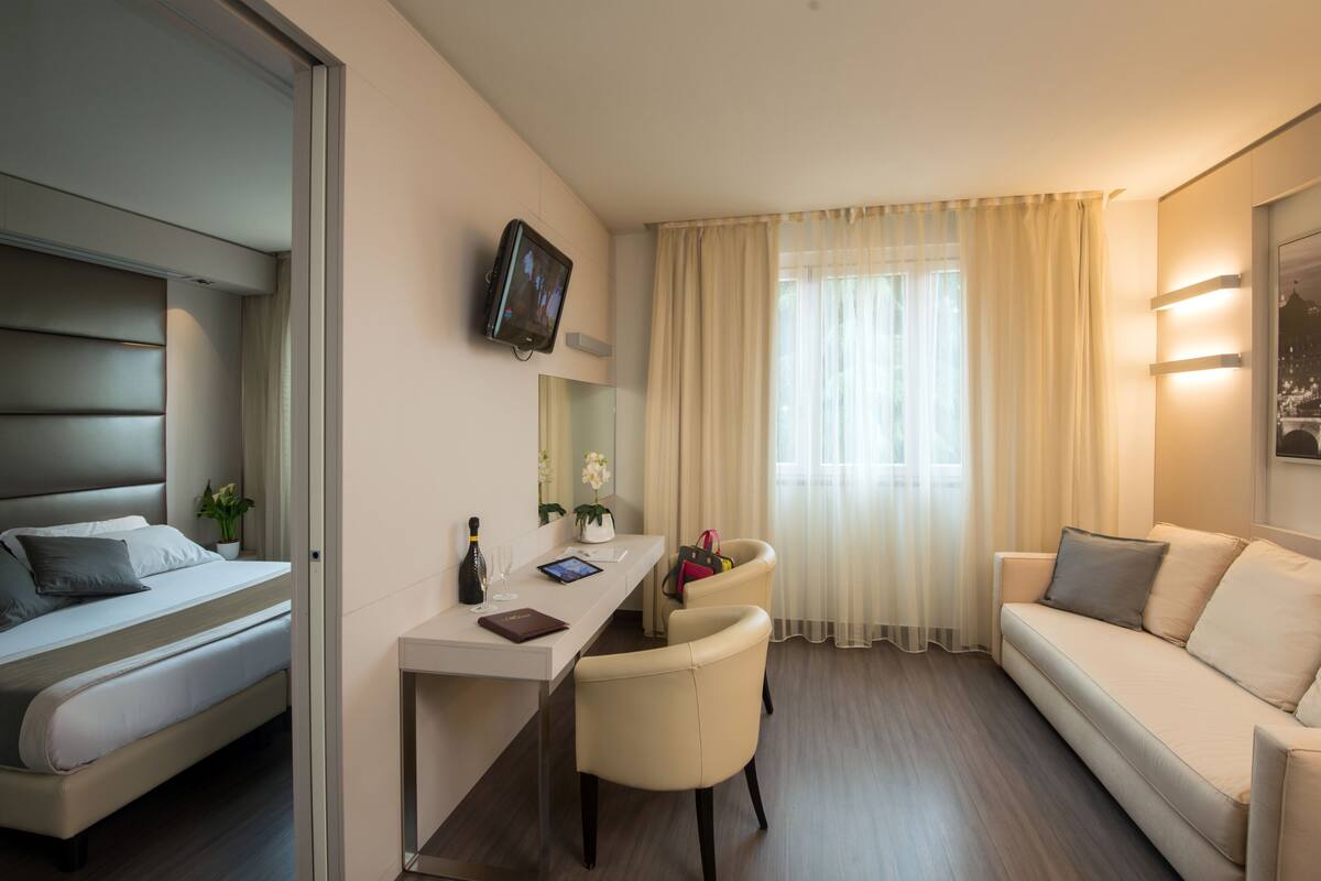 Ein modernes Hotelzimmer mit Bett, Schreibtisch, Stuhl, Sofa, Fernseher und einem Fenster mit Vorhängen.