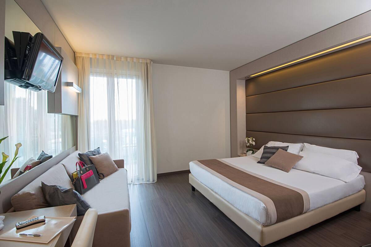 Ein modernes Hotelzimmer mit einem großen Bett, einer Couch, einem flachbildfernseher an der Wand und einem Schreibtisch mit Fernbedienung.
