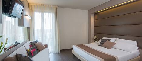 A modern hotel room with a large bed, a sofa, a flat-screen TV mounted on the wall, and a desk with a remote control.