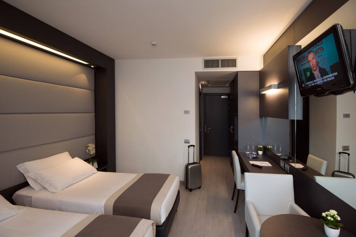 Ein Hotelzimmer mit zwei Betten, einem Schreibtisch, einem Stuhl, einem Fernseher und einem kleinen Tisch mit einer Pflanze.