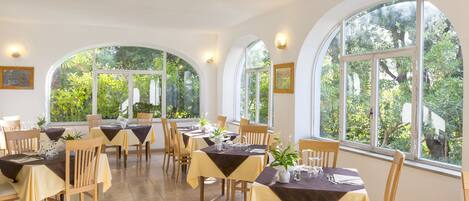 A dining area with tables set for a meal, large arched windows, and a view of greenery outside.