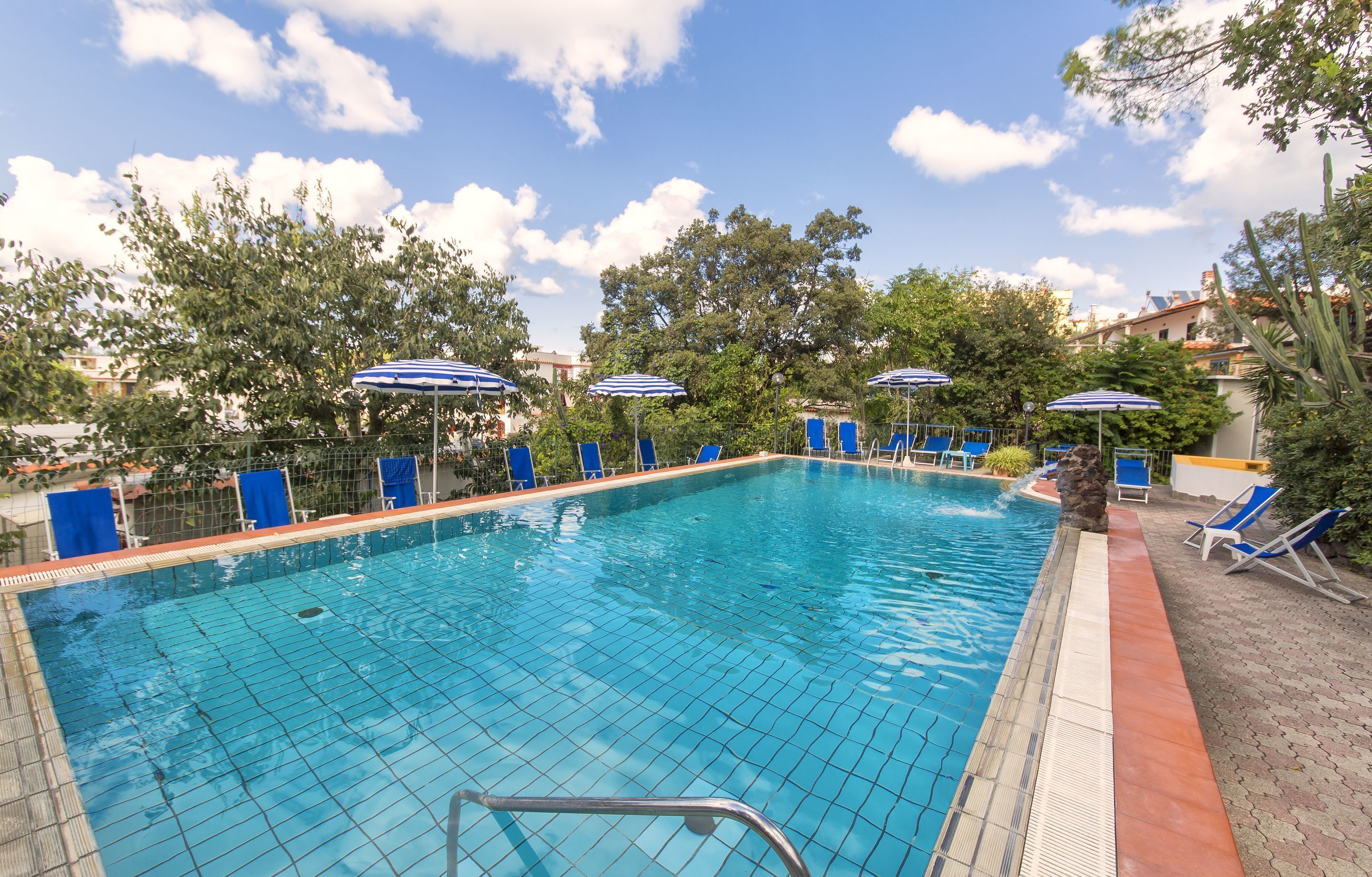 A swimming pool with blue chairs and umbrellas, surrounded by trees and buildings.