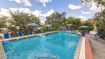 A swimming pool with blue chairs and umbrellas, surrounded by trees and buildings.