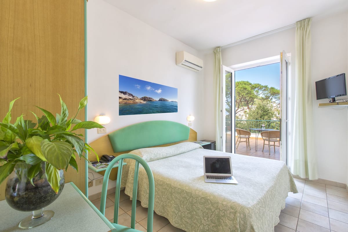 A hotel room with a bed, a laptop, a potted plant, a painting of a coastal scene, and a balcony with chairs.