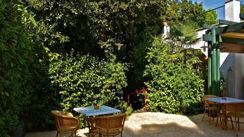 Un patio al aire libre con mobiliario de mimbre y una mesa, rodeado de follaje exuberante y una pared blanca.