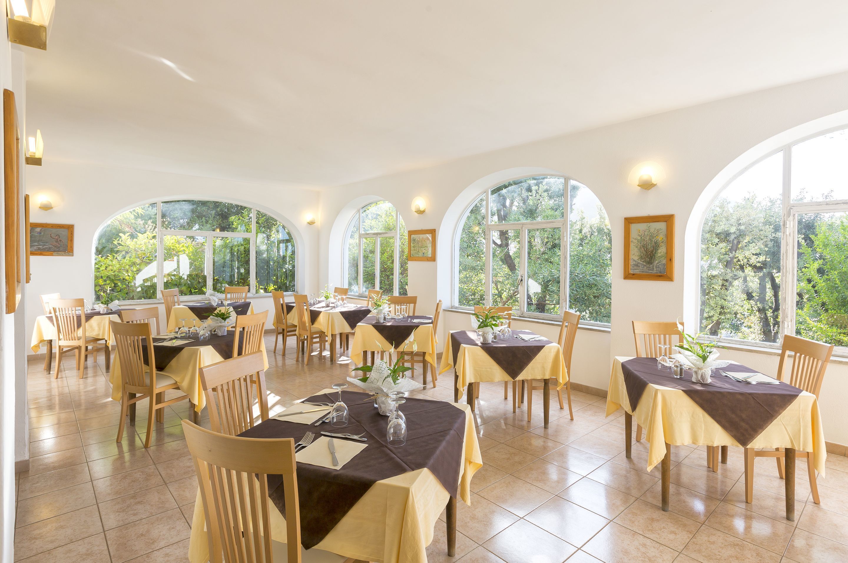 A dining area with multiple tables and chairs, large arched windows, and a view of greenery outside.