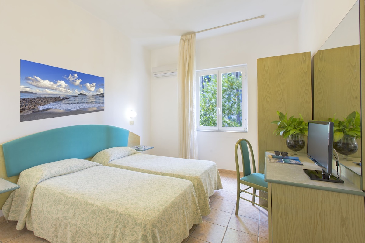 A room with two beds, a desk with a computer, a chair, a window with a view of greenery, and a painting of a beach scene.