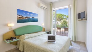 Ein Hotelzimmer mit Bett, einem Laptop auf dem Bett, einem Gemälde mit Meeresblick, einem Fernseher auf einem Schrank und einem Balkon mit Tisch und Stühlen.