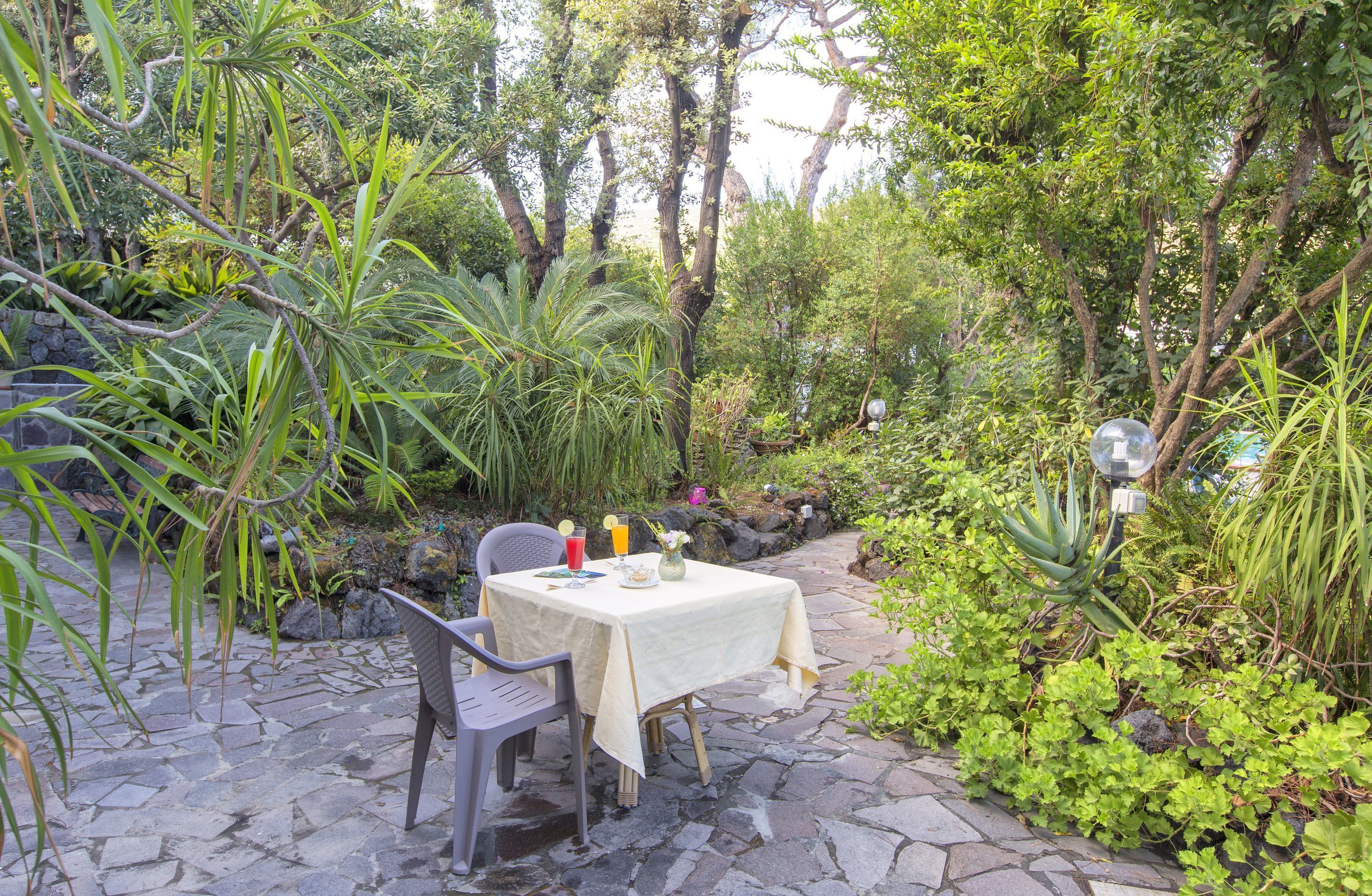 Zona pranzo all'aperto con tavolo per due, immersa nel verde e pavimentata in pietra.