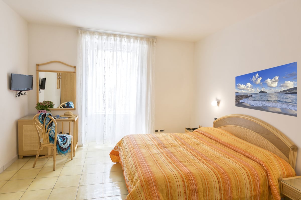 A bedroom with a bed, a desk, a chair, a television, and a window with curtains.