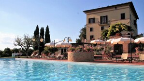 A pool area with lounge chairs and umbrellas, a stone planter with flowers, and a building with balconies in the background. - Villa Curina Resort (Castelnuovo Berardenga)