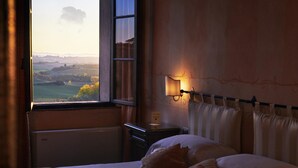 A hotel room with a bed, a nightstand, a lamp, and a view of a landscape through a window. - Villa Curina Resort (Castelnuovo Berardenga)